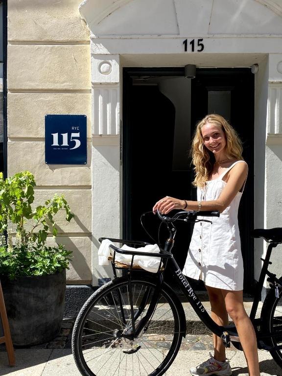 The entrance of Rye115 Hotel with its blue number sign and a guest standing with a hotel bicycle